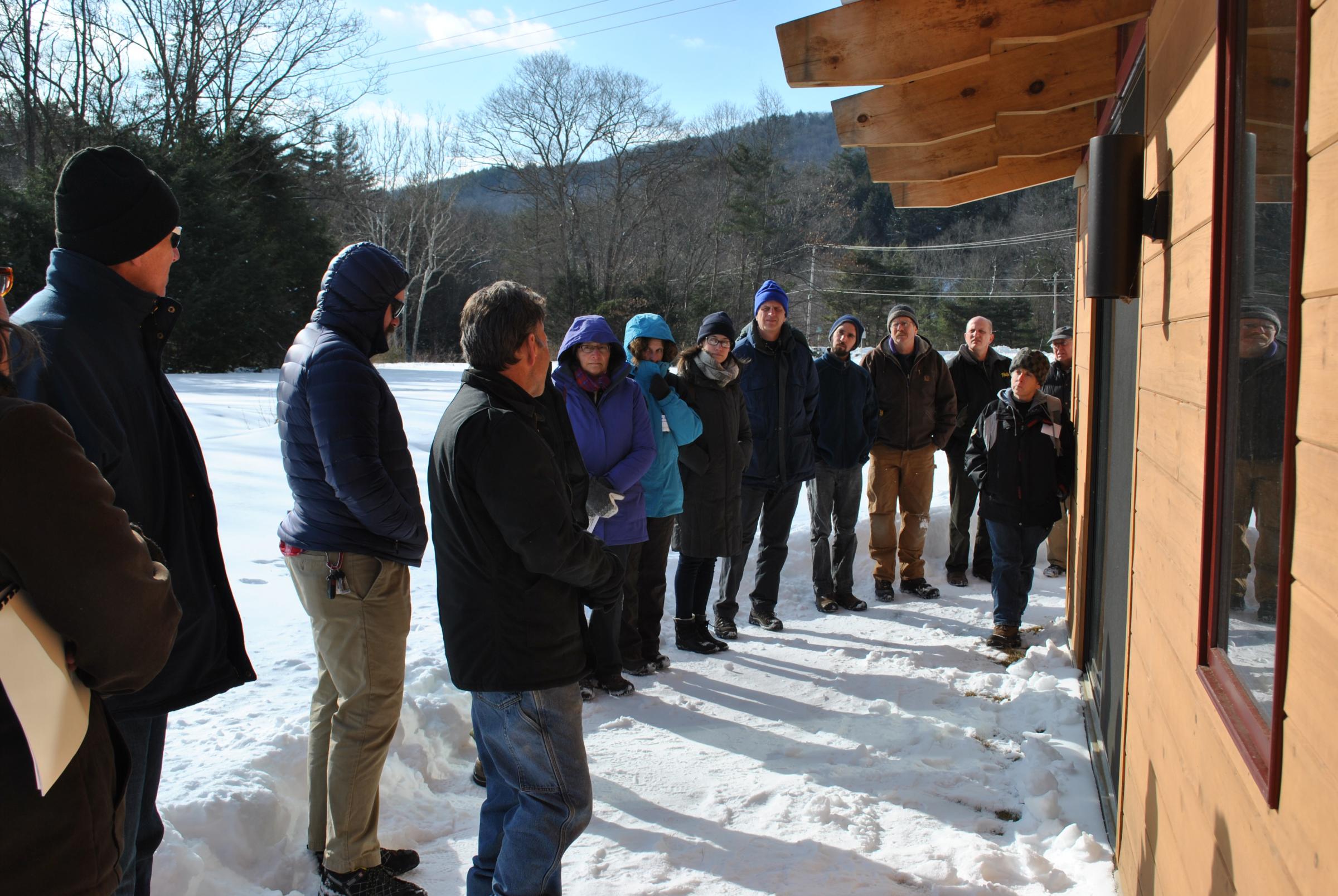 Group standing in a row next to a building during a BuildingEnergy Pro Tour (Caption: BuildingEnergy Pro Tour participants stand next to a building in Colrain, Mass. on Feb. 6, 2015.) 